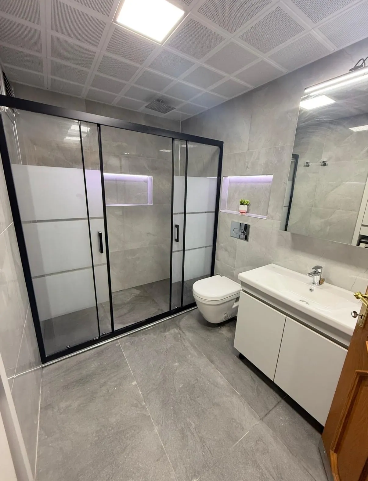 Siyah çerçeveli sürgülü duşakabin ve gri fayanslı banyo